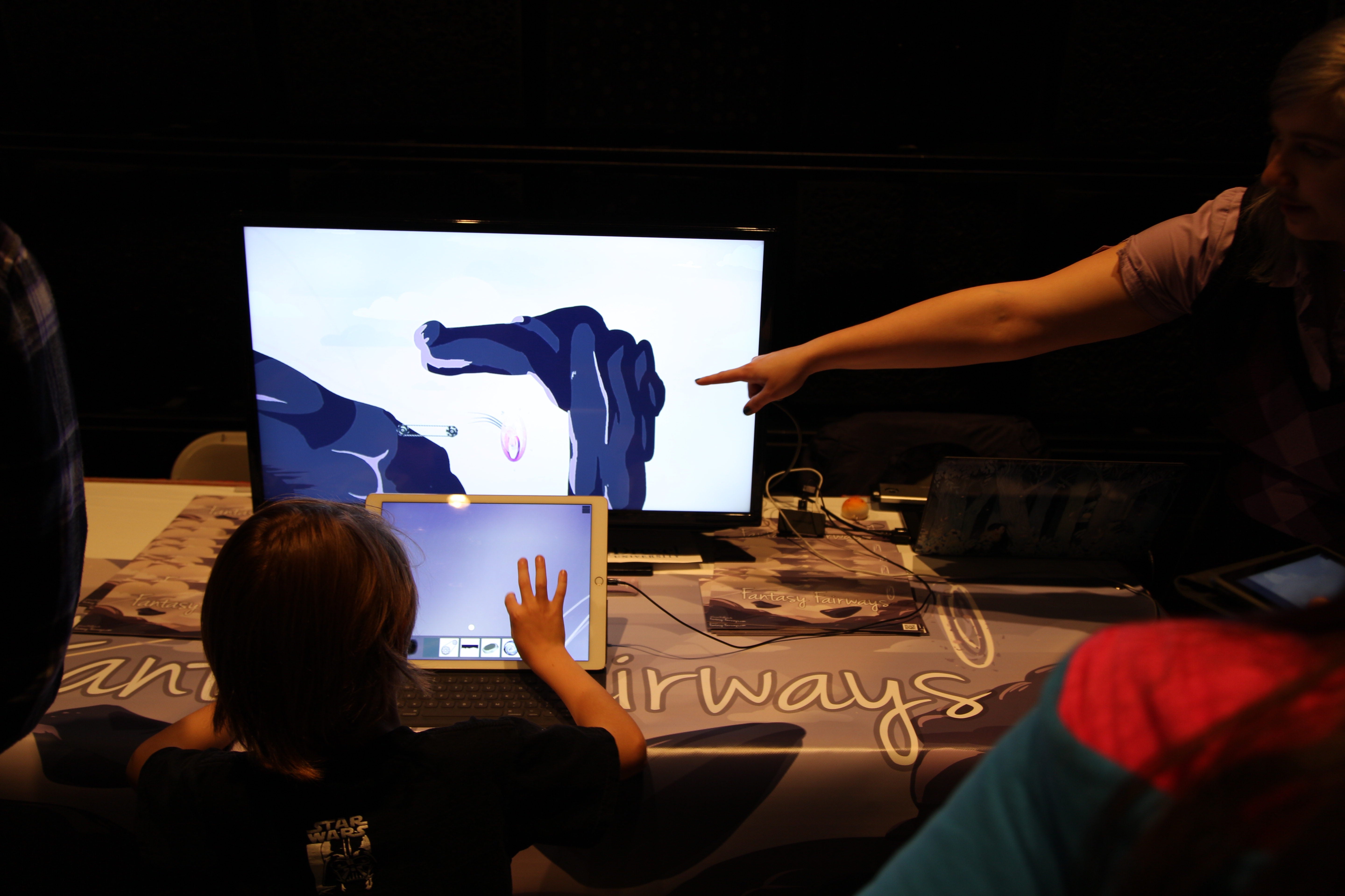 Student points at a screen as a child plays a game at GameFest 2016.
